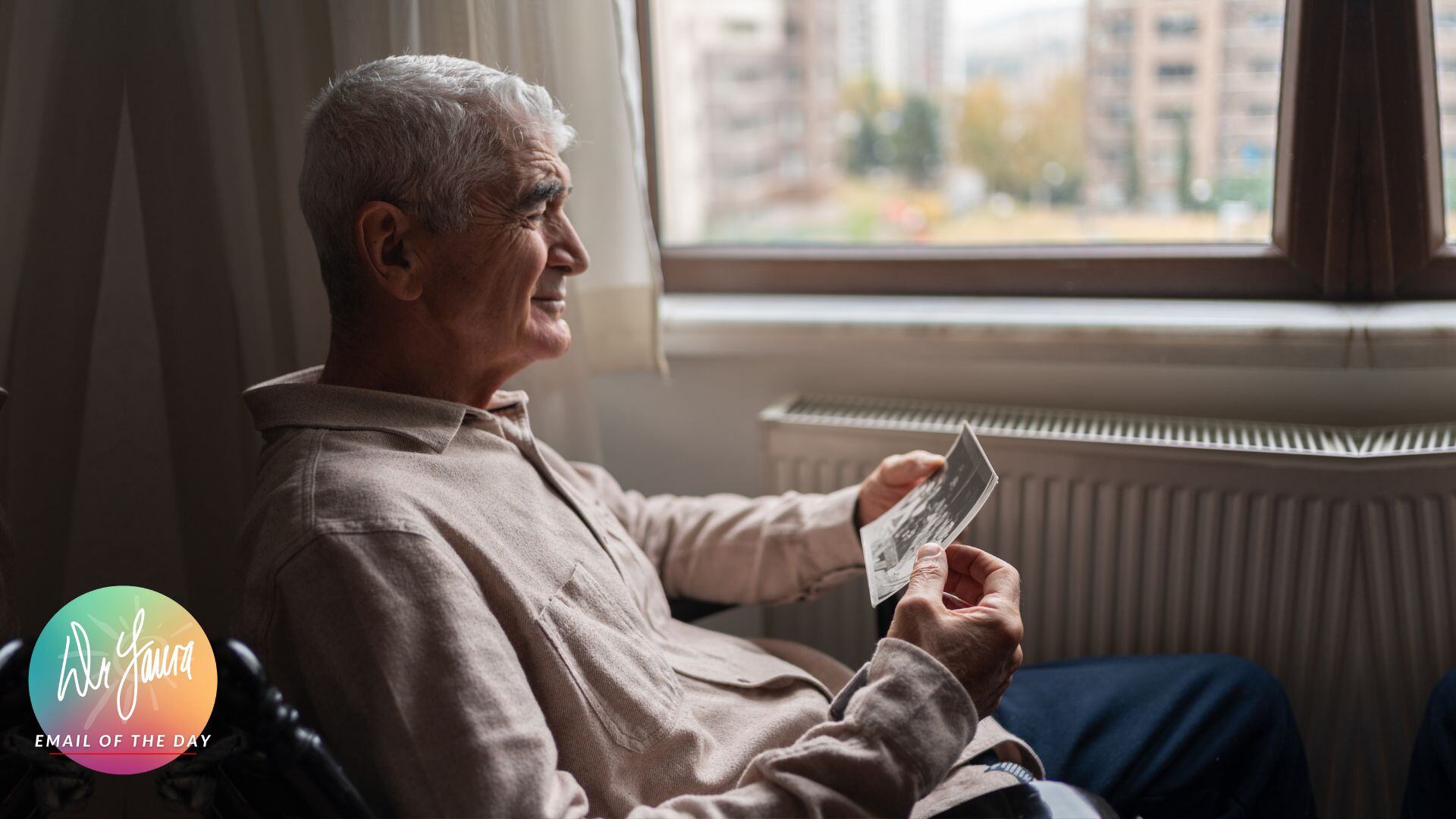 Older man sits on chair while looking out the window and holding a photograph